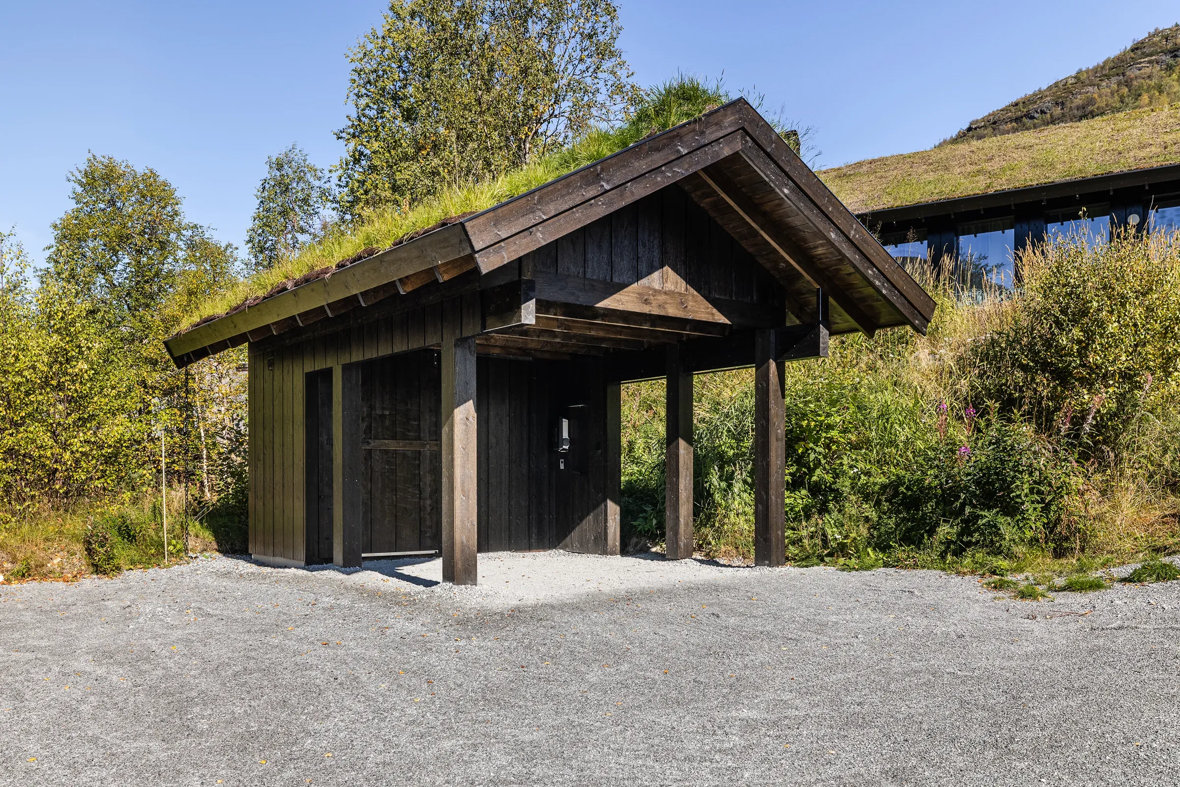 Carport med torvtak og mørk kledning, med hytten i bakgrunnen